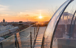 Sky Terrasse(Небо Терраса). Терраса с панорамным видом на город, ограждена стеклянными перилами. Справа расположена прозрачная конструкция с плетеной мебелью внутри, украшенной гирляндами. Закатное небо с ярким солнцем. Цвета: оранжевый, голубой, серый.