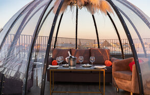 Sky Terrasse(Небо Терраса). Прозрачная геодезическая палатка на террасе. Внутри коричневая мягкая мебель: диван и кресло. Перед ними стол с белыми тарелками, бокалами и ведёрком для шампанского. Декор сверху из сухих растений. Железные перила на заднем плане.