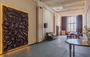 Chocolate Loft(Шоколад Лофт). Зал с видом на Финский залив. Просторное помещение с высоким потолком, белые стены, на одной из стен драпировка с подсветкой. Большое окно с темными шторами, стол и стулья, коричневый диван. Стена с колонками, деревянный круглый стол. Светодиодный светильник треугольной формы на потолке.