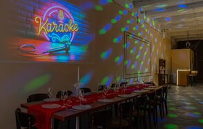 Chocolate Loft(Шоколад Лофт). Зал с видом на Финский залив. Пространство с длинным столом, покрытым красной дорожкой, сервировано бокалами и приборами. На кирпичной стене вывеска "Karaoke" с неоновыми микрофоном. Потолок высокий, с диско-освещением. Справа стойка бара, помещение в темных тонах.