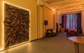 Chocolate Loft(Шоколад Лофт). Зал с видом на Финский залив. Пространство с высокими потолками, длинный стол с красными стульями, диван, деревянный круглый стол. Темные шторы, скульптурное панно с подсветкой, гирлянды на потолке. Светлые стены, темный пол, колонны. Освещение теплое, атмосферное.