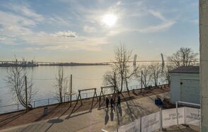 Chocolate Loft(Шоколад Лофт). Зал с видом на Финский залив. Пространство на набережной с видом на водоём и мост. Пешеходная дорожка с деревьями и уличным освещением. Светлое время суток, солнечно. Люди гуляют. Вдали видны портовые краны. Слева ограждение со знаками.