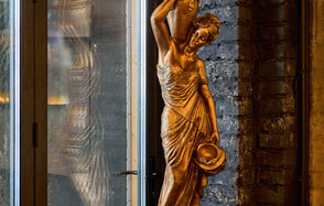 Tangiers Lounge(Танжер Лаундж) . Золотая статуя женщины в классическом стиле рядом с высоким стеклянным окном. Кирпичная стена тёмного цвета на заднем плане. Мягкое освещение подсвечивает детали скульптуры и текстуру стены.