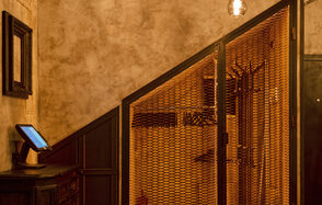 Tangiers Lounge(Танжер Лаундж) . Помещение с кирпичными стенами и деревянной отделкой. Под лестницей металлическая решетка; рядом темная деревянная мебель с экраном. Потолочный светильник с теплым освещением. Стены обшиты панелями, общей цвет — теплый, коричневый.