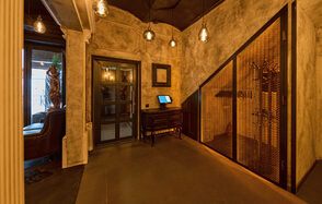 Tangiers Lounge(Танжер Лаундж) . Интерьерное пространство с приглушенным освещением, бетонными стенами и потолком. Тёмная дверь с стеклянной вставкой, небольшой стол с экраном. Деревянная решётчатая перегородка, подвесные светильники. Отделка из дерева и металла, нейтральные тона.