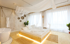 Пляжный лофт с галькой, открытой ванной, панорамными окнами. Просторное светлое помещение с кроватью на деревянном подиуме, подсветка под кроватью, белые шторы, подвешенный прозрачный балдахин. Ванна отдельно, круглая, белого цвета. Декоративные элементы на стене. Паркетный пол, две плетёные коврики.