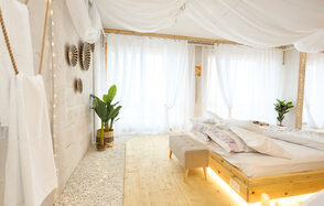 Пляжный лофт с галькой, открытой ванной, панорамными окнами. Светлое помещение с деревянным полом и гравийным покрытием. Большая кровать с декоративными подушками, деревянное изголовье. Прозрачные шторы, текстильный потолок. Декоративные растения, настенные украшения. Гардины пропускают естественный свет.