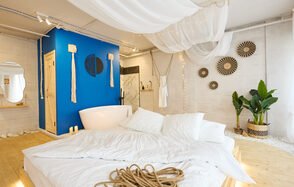 Пляжный лофт с галькой, открытой ванной, панорамными окнами. Просторная комната с белыми стенами, деревянным полом и ярко-синей акцентной стеной. Большая кровать с белым постельным бельём, рядом белая ванна. Растения в углу, потолок украшен тканью и светодиодными огнями. Декор: круглые элементы на стене.