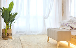 Пляжный лофт с галькой, открытой ванной, панорамными окнами. Светлое пространство с деревянным полом и белыми прозрачными занавесками. Слева стоят два плетёных горшка с зелёными растениями. Справа бежевый диван-бенч на деревянных ножках. На заднем плане видны постельные принадлежности.