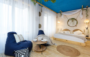 Лофт Бали с фото зоной на 23 этаже с видом на Москва-реку. Спальня с деревянным полом и синим потолком, кровать с белым постельным бельём, два кресла-мешка, круглый стол, подвесные светильники, декоративные растения и круглый ковёр. Белые занавески на окне, бежевые и черные подушки.