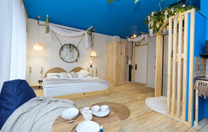 Лофт Бали с фото зоной на 23 этаже с видом на Москва-реку. Спальня с двуспальной кроватью, деревянными панелями и голубым потолком. Декор из растений, светильники, круглый стол с посудой, кресло-мешок. Светлые деревянные полы, стены декорированы тканями и венком. Гардероб с открытыми полками.