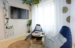 Лофт Бали с фото зоной на 23 этаже с видом на Москва-реку. Современное пространство с деревянным полом, двумя синими креслами-мешками, круглым кофейным столиком с белыми тарелками и чашками. Белая штора на окне, телевизор на стене, декоративные элементы из растений и труб, мягкое освещение.