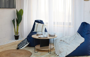 Лофт Бали с фото зоной на 23 этаже с видом на Москва-реку. Современная гостиная с двумя синими креслами-мешками, круглым деревянным столом с посудой, растением в горшке, светлыми шторами, круглым ковриком и деревянным полом. Белая кирпичная стена с телевизором, декор в нейтральных тонах.