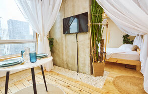 Тропический лофт с бунгало, баром и французским балконом. Светлое помещение с деревянным полом и тканевым навесом. В центре бетонная стена с телевизором. Слева стол с посудой, справа кровать. У стены большие окна, декорированы растения. Под ногами ковровая дорожка и белая галька.