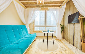 Тропический лофт с бунгало, баром и французским балконом. Просторная комната с деревянным полом и потолком. Голубой диван у стены, напротив — круглый стол с черными ножками. Белые занавески на окнах и вокруг дивана. На стене телевизор, рядом растение. Светлая, минималистичная обстановка.