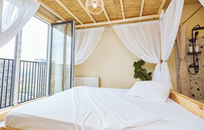 Тропический лофт с бунгало, баром и французским балконом. Спальня с двуспальной кроватью, белым постельным бельем и балдахином. Большое окно и стеклянная дверь ведут на балкон с черными перилами. Натуральный потолок с деревянными балками. На стене радиатор. В углу зелёное растение. Светлые занавеси.