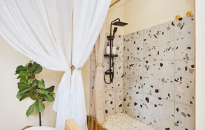 Тропический лофт с бунгало, баром и французским балконом. Душевая зона с терраццо плиткой на стене, черный душ, светлые ткани, бежевые стены, растение в горшке, деревянные элементы, стеклянные бутылочки на полке, белый текстиль.