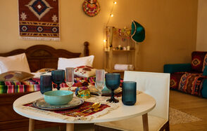 ⁠Лофт в Мексиканском стиле для костюмированных вечеринок. Гостиная с деревянным столом, белыми стульями и сервировкой из цветных бокалов и тарелок. На стене текстильный декор, рядом кровать с разноцветным покрывалом, в углу диван с ярким пледом. Теплое освещение и деревянный пол.