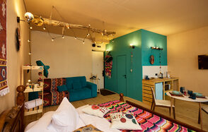 ⁠Лофт в Мексиканском стиле для костюмированных вечеринок. Гостиная с кроватью, диваном, столом и стульями. На голубой стене декор и полка с посудой. Деревянная мебель, яркие ткани с узорами. Верхнее и гирляндное освещение. Деревянный пол, стены светлые. Бежевые и голубые цвета. Телевизор на стене.