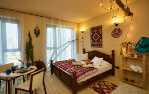 ⁠Лофт в Мексиканском стиле для костюмированных вечеринок. Просторная комната с деревянным полом, большая кровать с текстилем ярких цветов, рядом деревянный стол и два стула. На стене тканевые декорации, полка с декором. Окна в пол, через которые проникает свет. Потолочный декор с лампами, светлый текстиль на окнах.