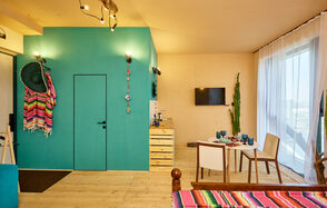 ⁠Лофт в Мексиканском стиле для костюмированных вечеринок. Комната с деревянным полом, бирюзовой стеной и декором. Белые и деревянные стулья вокруг стола с посудой. Потолочные светильники, телевизор на стене, зелёное растение у окна с белыми шторами. Цветные пледы на стуле и стене.