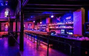 Castle Concert Hall(Концертный зал Замок). Барное пространство с длинной стойкой и высокими стульями, освещение фиолетового и розового оттенков. На заднем плане стеллажи с бутылками, неоновые элементы на стенах. Металлические и деревянные элементы, открытые трубы и балки.