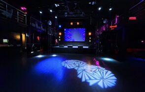 Castle Concert Hall(Концертный зал Замок). Ночной клуб или концертный зал с темным интерьером, большой экран на сцене, разноцветные прожекторы и световые эффекты на полу, металлические перила, второй ярус, черный пол и стены, современные осветительные приборы, отсутствуют посетители.