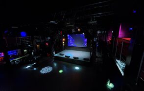 Castle Concert Hall(Концертный зал Замок). Темное пространство с большой сценой, экраном и разноцветными световыми эффектами. Металлические перила и конструкции, отражающие поверхности. На стенах подсветка красного и синего цветов. Светящиеся кружки на полу. Балконы и лестницы по краям.