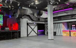 PRAVDA(Правда). Просторный зал на 800 гостей. Просторное помещение с бетонным полом и колонной в центре. Серые стены с черными элементами. Площадка с оборудованием и красной подсветкой. Барная стойка с фиолетовой и розовой подсветкой. Потолочные светильники и аудиосистема.