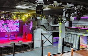 PRAVDA(Правда). Просторный зал на 800 гостей. Клубное помещение с большой сценой, оснащённой экранами и звуковым оборудованием. Верхний этаж с балюстрадой и зонами для сидения. Освещение разноцветное, включая прожекторы. Стены серые, полы тёмные. Потолок в чёрных конструкциях. Отсутствие людей.