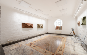 Светлый Лофт. Прямоугольное помещение с белыми кирпичными стенами и мраморным полом, украшенное настенными инсталляциями. Арочное окно с круглым проемом, прозрачные стулья, осветительные трековые светильники, бежево-коричневый ковер, деревянный штатив.