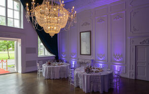 Лофт КОСМОС с летней верандой. Большая комната с роскошной люстрой, двумя круглыми столами с белыми скатертями и прозрачными стульями. Декоративные панели на стенах, одно зеркало. Фиолетовая подсветка, темно-синие шторы, деревянный пол, высокие окна и двери.