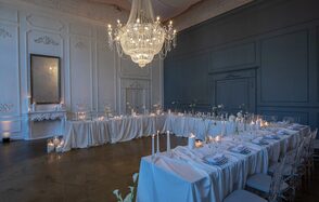 Лофт КОСМОС с летней верандой. Праздничное пространство с длинными столами в форме буквы «Г», покрытыми белыми скатертями и сервировочными наборами. Стулья прозрачные, вокруг свечи. Центральный элемент — большой хрустальный светильник. Стены белого и серого цветов с панелями.