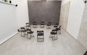 Интер-Фото. Просторное помещение с белыми стенами и светлым плиточным полом. В центре расставлены черные складные стулья по кругу. Стены декорированы серыми и белыми текстурными панелями. Есть рулон белого фона, установленного на стойке.