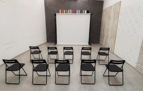 Интер-Фото. Пространство с 12 черными стульями, установленными перед экраном на штативе. Пол бетонный, стены светлые с текстурой. Потолок не виден, сбоку стена имеет круглый узор, рядом разноцветные цилиндры на стойке.