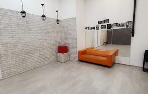 Интер-Фото. Просторная комната с белым кирпичным декором стен. На полу светлая плитка. Оранжевый диван расположен перед зеркальной стеной. Подвесные черные светильники. На стене черно-белые фотографии. Белая квадратная корзина и красный куб.