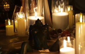 Лофт Поле. Уютный зал для мероприятий. Свечи в прозрачных стаканах и большие белые свечи на столе, украшенном тканью. Темная фигурка декора, вокруг небольшие камни. Освещение теплое и мягкое, создающее атмосферу уюта и интимности.