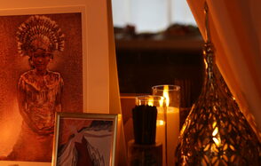 Лофт Поле. Уютный зал для мероприятий. Картина с изображением человека в головном уборе, установлена на столе рядом с двумя свечами в стеклянных подсвечниках. Рядом декоративный металлический элемент с узорами. Освещение мягкое и теплое.