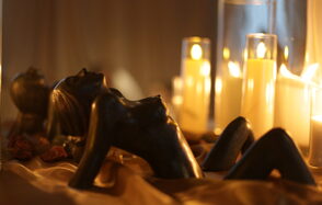 Лофт Поле. Уютный зал для мероприятий. Свечи в стеклянных подсвечниках горят на фоне. Темная скульптура фигуры человека лежит на тканевой поверхности при мягком освещении. Цветовая гамма теплая, преобладают золотистые и коричневые оттенки.