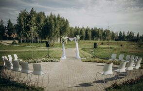 Эко-отель "Джао Да!Ча". Шатер-Сфера. Открытое пространство с кирпичной дорожкой, окружённой газоном. Арка, украшенная белым текстилем и цветами. По бокам арки установлены колонки на стойках. По обе стороны дорожки расположены белые стулья в два ряда, образуя полукруг. Позади деревья.