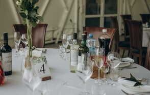 Эко-отель "Джао Да!Ча". Шатер-Сфера. Круглый стол, покрытый белой скатертью, сервирован бокалами и бутылками напитков, включая вино и газировку. На столе декоративная композиция из зелени и карточка с номером 4. Вокруг деревянные стулья.