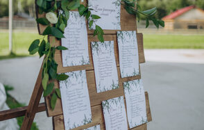 Эко-отель "Джао Да!Ча". Шатер-Сфера. Деревянная подставка с планом рассадки гостей оформлена белыми карточками с текстом и украшена зелеными листьями и цветами. На заднем плане виден участок земли с зелёной травой и зданием.