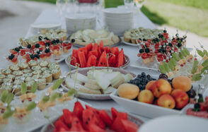 Эко-отель "Джао Да!Ча". Шатер-Сфера. Стол с тарелками, заполненными фруктами, такими как арбузы, дыня, абрикосы и виноград. Также представлены десерты и закуски на шпажках. Посуда белая, вокруг видна зелёная трава и расставлены стаканы.