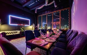 Лофт Овертон. Темное помещение с длинным столом, сервированным тарелками и бокалами, окруженным мягкими стульями. На стене неоновая декоративная панель. Большие окна в раме выходят на ночной город. Диван стоит у стены. Светодиодные гирлянды на потолке.