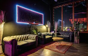 Лофт Овертон. Лофт-пространство с черными стенами и потолком, три серых дивана, низкий деревянный стол, ковёр с узором, неоновая подсветка на стене, большие окна, деревянный пол. Декор из растений в углах, стойка с зеркалом, стена с графическим узором в неоновом свете.