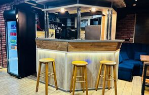 BULL LOFT(Лофт Бык) . Барная стойка с деревянной обивкой и тремя высокими стульями, подсветка снизу, фонарики сверху. В глубине shelving с бутылками и стаканами. Напольное покрытие светлое. В углу черный холодильник с напитками. Справа диван темного цвета.