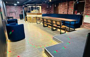 BULL LOFT(Лофт Бык) . Кирпичные стены, длинный деревянный стол с лавками и мягкими креслами, кухня с барными стульями в углу, паркетный пол, мягкий ковролин на возвышении, яркое точечное освещение, разноцветные световые эффекты на полу, черный потолок с подвесными светильниками.