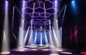 Studio Hall(Студио Холл). Большое пространство в индустриальном стиле, с кирпичными стенами и высокими потолками. Центральные светильники в виде гексагонов, светодиодное освещение разных цветов. Пустая сцена с направленной светотехникой, пол темный.