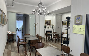 Арт Холл. Просторное помещение с деревянными полами, расставленными деревянными столами и стульями. На стенах картины. Две люстры с лампами. Стеклянные раздвижные двери ведут в другую комнату. Есть камин, полки с декором и стойка для одежды.