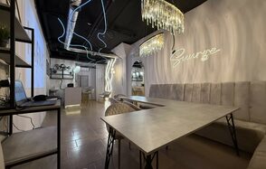 Лофт SUNRISE(Восход Солнца). Просторное помещение на 15 гостей. Прямоугольное помещение с длинным столом и стульями. Вдоль стены мягкие диваны. Потолочные светильники с хрустальными элементами. Светлая плитка на полу. На стене неоновая надпись. Открытые полки с растениями и техникой. Темный потолок с вентиляцией.